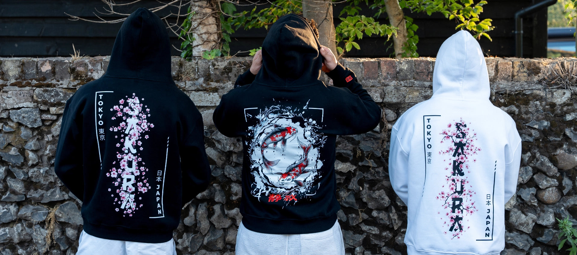 ArcaLoot Sakura and Serenity black and white hoodies worn by three models against a cobblestone wall. Featuring Japanese cherry blossom and koi fish designs.
